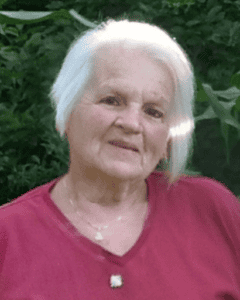 Photo of Denise before her passing. Denise wearing a red shirt sitting in front of green foliage. She has white chin length hair.